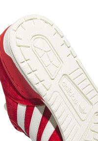 Zapatilla de gamuza roja con rayas de cuero blanco y suela de goma blanca texturizada. Presenta el logo de Adidas en el lateral y un patrón de banda distintivo.