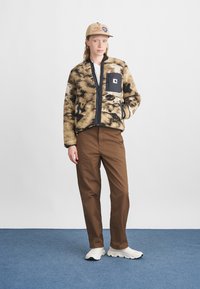 Chaqueta de felpa con patrón marrón y negro, con cremallera y bolsillos laterales, combinada con pantalones marrones y zapatillas blancas. Gorra marrón con detalle de logo.