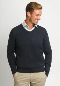 Marineblauwe v-hals trui van zacht materiaal, met lange mouwen en een geribbelde zoom, gedragen over een gekruiste lichtgeruite blouse.