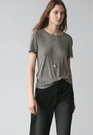 Young woman wearing a short-sleeve gray top with black lace trim, black high-waisted jeans, and a long gold pendant necklace.