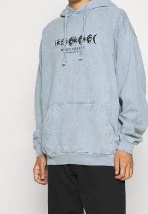 Blaugrauer Hoodie mit schwarzer Mondphasen-Grafik und Text "Moon Phases AOTIKO Society,", getragen mit schwarzer Hose, zeigt Torso und Arme.