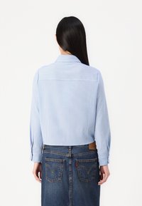 Camicia a righe blu chiaro con colletto, polsini a bottoni e retro accorciato. Abbinata a una gonna di denim con tasche posteriori e una toppa con logo.