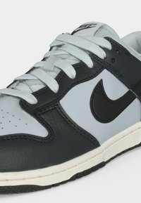 Μαύρο και γκρι δερμάτινο αθλητικό παπούτσι Nike με λευκά κορδόνια, διάτρητο μπροστινό μέρος και λογότυπο στο πλάι και στη γλώσσα, πλάγια μπροστινή άποψη.