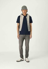 Mladi muškarac nosi sivi šešir, tamnoplavu polo košulju, svijetlosivu prsluk bez rukava preko ramena, sive traperice i bijele tenisice s crnim trakama.