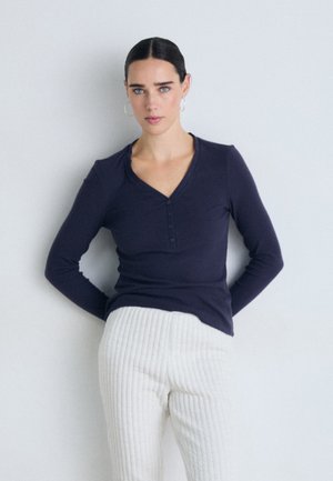 Navy long-sleeved top with a V-neck and three-button placket, paired with cream ribbed trousers featuring a textured pattern.