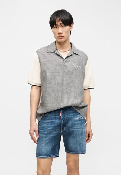 Jeune homme portant une chemise beige à manches courtes à carreaux avec un col et un short en jean bleu, debout devant un fond blanc uni.