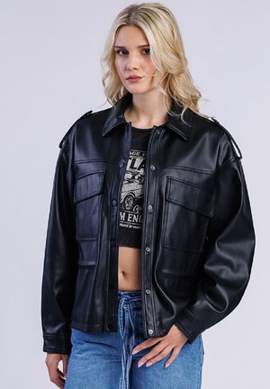 Blonde Frau trägt eine schwarze Kunstlederjacke über einem kurzen Graphic-Top und blauen Jeans, steht vor einem schlichten Hintergrund.