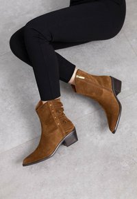 Botas de tobillo de ante marrón con punta afilada, detalles de flecos, tachuelas decorativas y una cremallera lateral. El tacón apilado añade estabilidad.