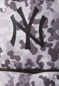 Zaino con motivo camouflage in tonalità di grigio, con un grande logo nero NY. Presenta un tessuto resistente e una tasca con zip nella parte inferiore.