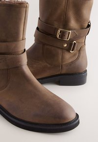 Bottines en cuir marron avec un bout arrondi, un détail de boucle métallique et une finition texturée. Semelle plate noire avec surpiqûres contrastantes.