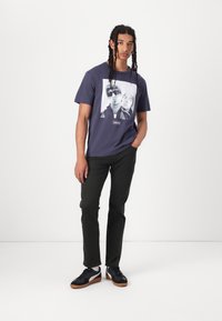 Navy graphic t-shirt featuring a black-and-white photo print, paired with dark green pants and black sneakers with gum soles.