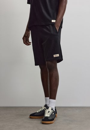 Bărbat purtând pantaloni scurți negri texturiți și tricou asortat, cu șosete albe și adidași negri, stând lângă un perete gri.