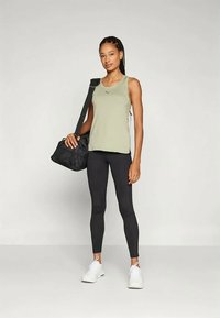 Kvinne i grønn ermeløs topp, svarte leggins, hvite joggesko, som bærer en svart treningsbag over den ene skulderen, stående mot en ensfarget bakgrunn.