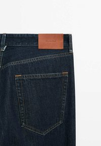 Vue de dos d'un jean en denim bleu foncé montrant une seule poche arrière et une étiquette de marque en cuir marron sur la ceinture.