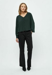 Blusa verde scuro a maniche lunghe con scollatura a V, bottoni frontali e vestibilità rilassata, abbinata a pantaloni neri a gamba dritta e scarpe nere a punta.