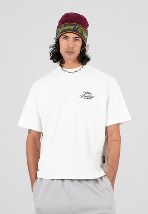 Weißes T-Shirt mit kurzen Ärmeln und rundem Halsausschnitt, das ein kleines schwarzes gesticktes Logo auf der Brust zeigt. Kombiniert mit grauen Hosen und einer burgunderfarbenen Strickmütze.