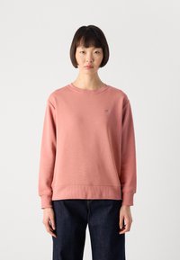 Woman with short dark hair wearing a pink crewneck sweatshirt and dark jeans, standing against a plain white background.