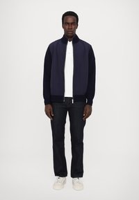 Navy bomber jacket with ribbed cuffs, zippered front, and side pockets, paired with a white polo and dark denim jeans, worn with white sneakers.