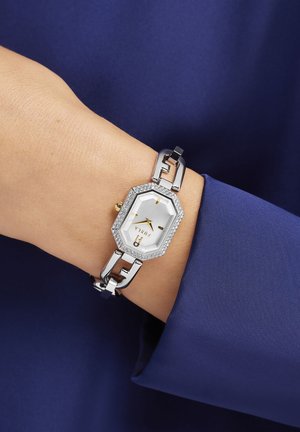 Montre en métal argenté avec un cadran octogonal, un cadran blanc, des accents dorés et des embellissements en cristal sur la lunette. Portée sur un poignet recouvert de tissu bleu.