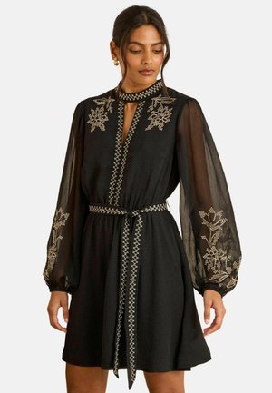 Sort kjole med gennemsigtige lange ærmer, broderede blomsterdetaljer, nøglehulshalsudskæring og et stofbælte, der fremhæver en mønstret kant.