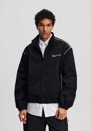Veste noire à fermeture éclair avec des poignets et un ourlet en bas élastiques, présentant des passepoils blancs contrastants et un logo sur la zone de la poitrine gauche.