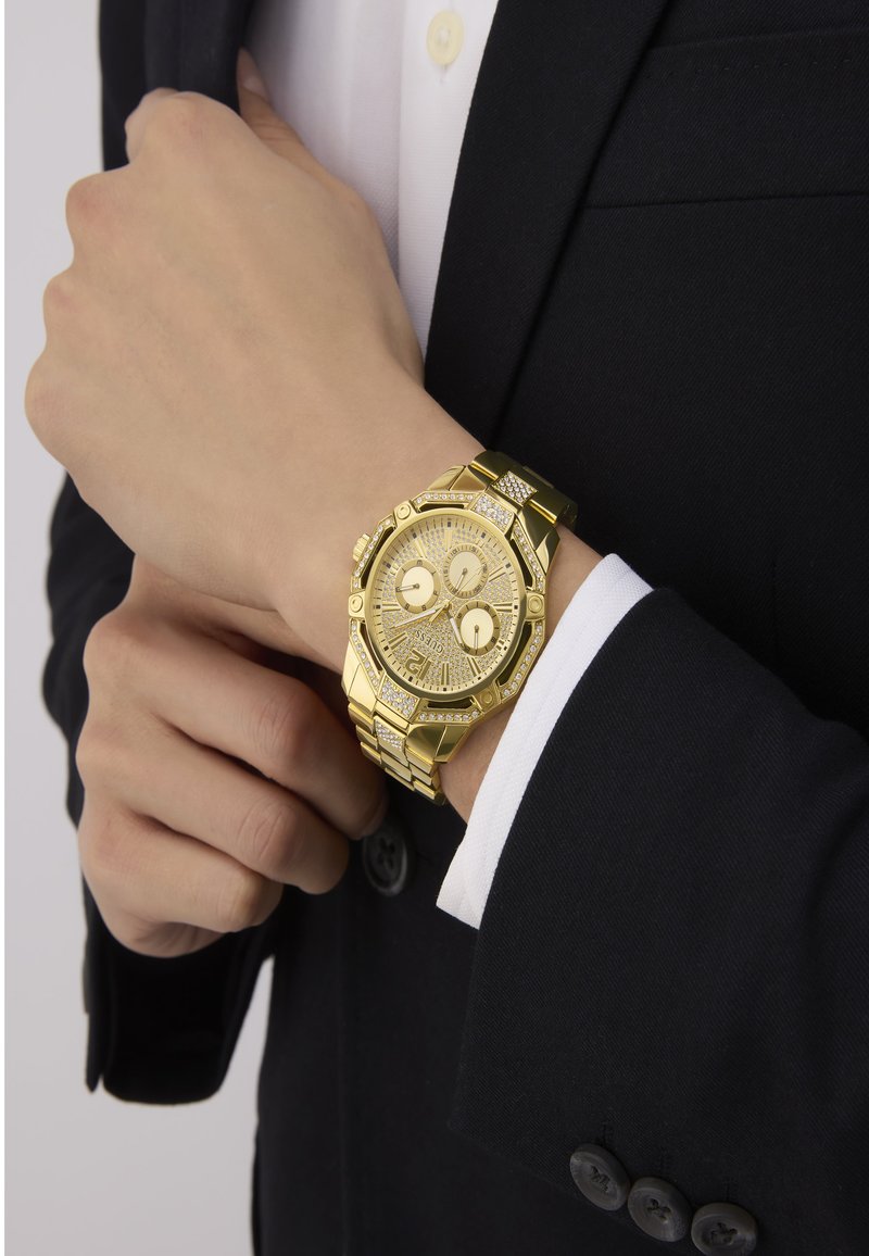 Goldfarbene analoge Armbanduhr mit Chronographenfunktionen, strukturiertem Ziffernblatt, Kristallakzenten und einem goldfarbenen Gliederarmband, getragen über einem dunklen Sakko.