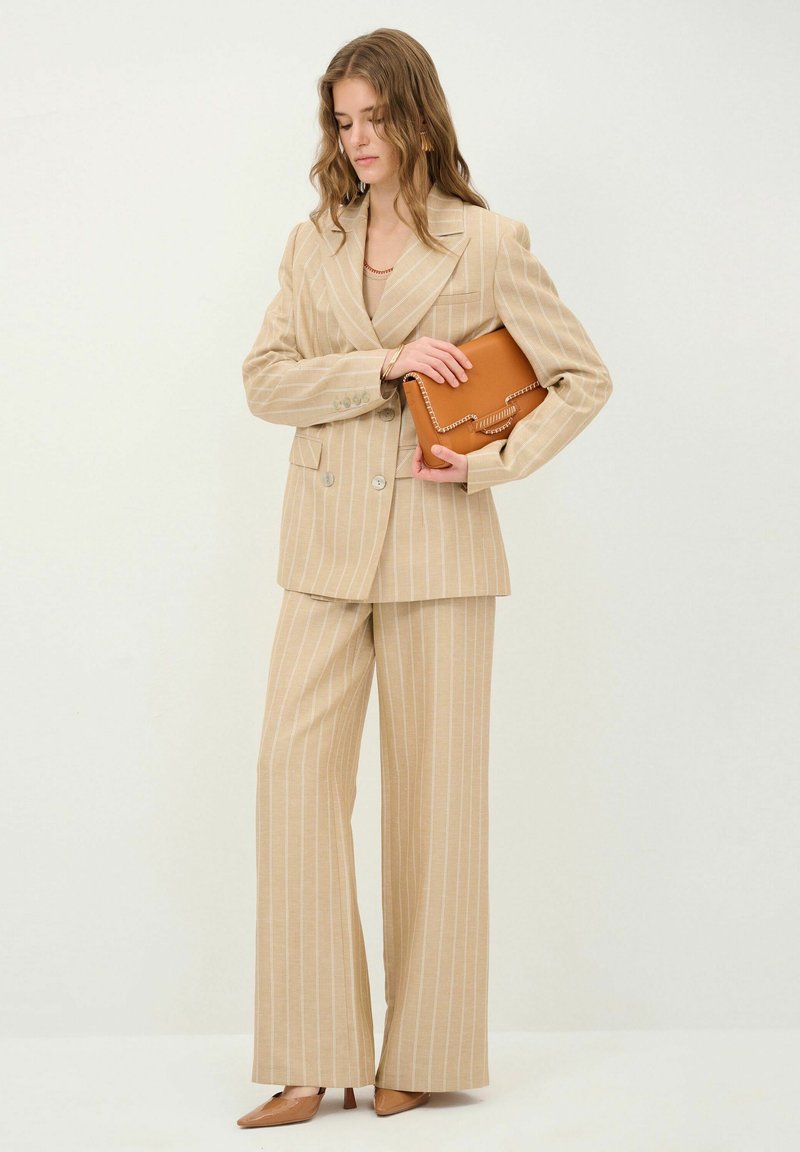 Beige stribet jakkesæt lavet af blødt stof, med brede bukser og en dobbeltknappet blazer. Komplementeret med en brun clutch.