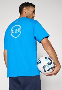 Blå bomulds T-shirt med et cirkulært "ATLETİ" logo på ryggen. Modellen holder en hvid fodbold med blå og sorte accenter.
