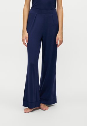 Femme portant un pantalon large taille haute bleu marine, debout pieds nus devant un fond clair uni.