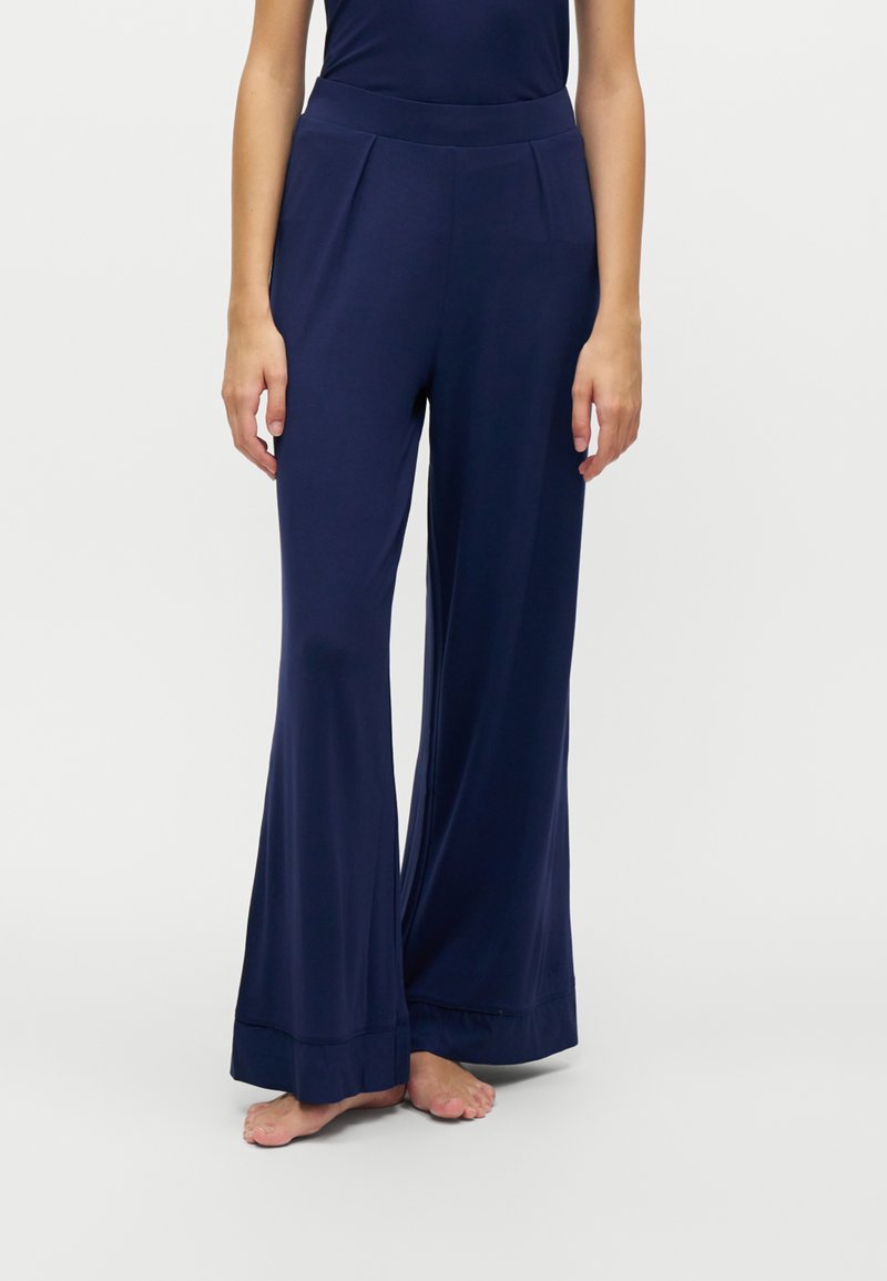 Femme portant un pantalon large taille haute bleu marine, debout pieds nus devant un fond clair uni.