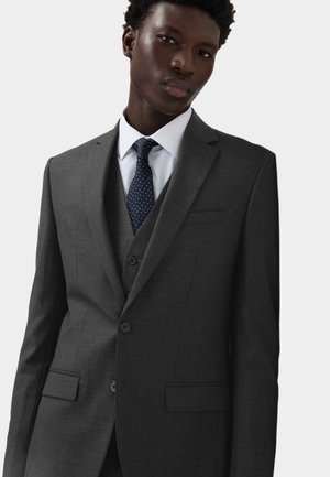 Costume trois pièces gris foncé avec revers crantés, veste à simple boutonnage, deux boutons, et une coupe ajustée, associé à une chemise blanche et une cravate bleu marine à pois.