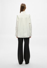 Fehér ing magas nyakkal, hátul hajtással és gombos mandzsettával, széles fekete nadrággal párosítva. Sima textúra és laza szabás.