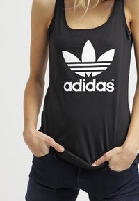 Svart ärmlös linne med en stor vit Adidas-logotyp och det karakteristiska tre-stripsdesignet. Mjuk tygkvalitet med en figurnära form.