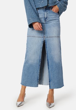 Kvinde iført en lyseblå denim midi-nederdel med frontsplit, sølvfarvede spidse hæle og en tekstureret blå jakke med lange ærmer.
