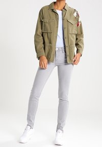 Olivově zelená vojenská bunda se předními kapsami a květinovou výšivkou na rameni, v kombinaci s světle šedými skinny džíny a bílými teniskami.