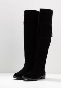 Botas de ante negras por encima de la rodilla con tacón plano, que presentan una textura suave y un zipper lateral para facilitar su uso. Diseñadas para un ajuste ceñido.