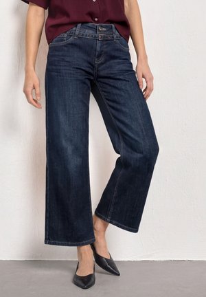 Kvinde iført mørkeblå vide jeans, sorte spidse flade sko og en bordeaux top, stående mod en lys tekstureret væg.