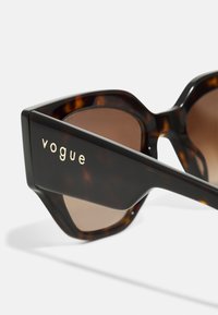 Braune Schildpatt-Sonnenbrille mit schrägen Rahmen, die ein goldenes "Vogue"-Logo an der Seite und braun getönte Gläser besitzt.