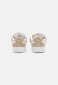 Baskets en suède beige avec une semelle en caoutchouc blanc, comportant des accents en cuir blanc et un petit logo jaune sur le talon.