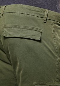 Bawełniane spodnie w oliwkowej zieleni z tylną kieszenią, z płaskimi szwami i gładką fakturą. Widoczne szlufki na pasek w talii.
