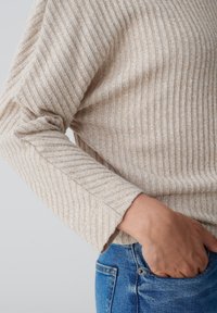 Hellbeige gerippter Pullover mit langen Ärmeln, der subtile silberne Fäden aufweist, kombiniert mit blauen Jeans. Texturierte Stoffdetails sichtbar.