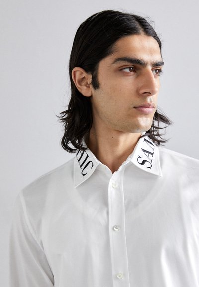 White button-up shirt with a structured collar featuring black embroidered text. The fabric appears smooth with a slight sheen.