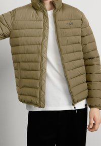 Olivgrön vadderad jacka med quiltad design, hög krage och dragkedja framtill. Har en sidoficka och en diskret logotyp på bröstet.