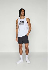 Joven con el pelo trenzado que lleva una camiseta sin mangas blanca de "JACK & JONES", pantalones cortos negros, calcetines blancos y zapatillas blancas, de pie con una mano en el bolsillo.