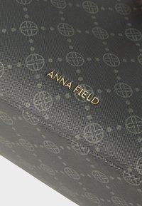 Tissu sombre texturé avec un motif circulaire répétitif et le lettrage doré du logo "ANNA FIELD" dans le coin d’un sac ou accessoire.