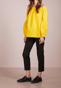 Žlutý top s odhalenými rameny a nabíranými rukávy, doplněný černými zúženými kalhotami a černými teniskami. Hladká textura a jednobarevné provedení.