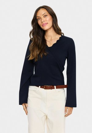 Vrouw met lang bruin haar, gekleed in een marineblauwe v-hals trui en Beigekleurige broek met een bruine leren riem, staand tegen een effen achtergrond.