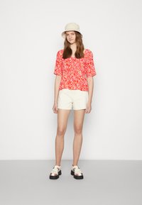 Blusa de manga curta vermelha com padrão floral, calções brancos e sandálias grossas. Um chapéu de balde bege complementa as cores e o design casual do conjunto.