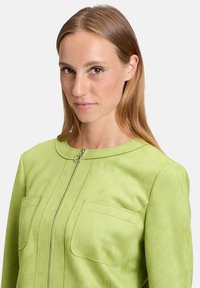 Zielona zamszowa kurtka z okrągłym dekoltem, zamkiem błyskawicznym z przodu i dwiema kieszeniami na piersiach. Tkanina ma gładką fakturę i subtelny połysk.