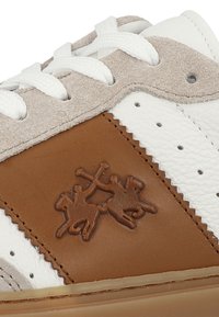 Białe skórzane sneakersy z brązowymi skórzanymi akcentami, zamszowym językiem, perforowanymi detalami i wytłoczonym wzorem po boku.